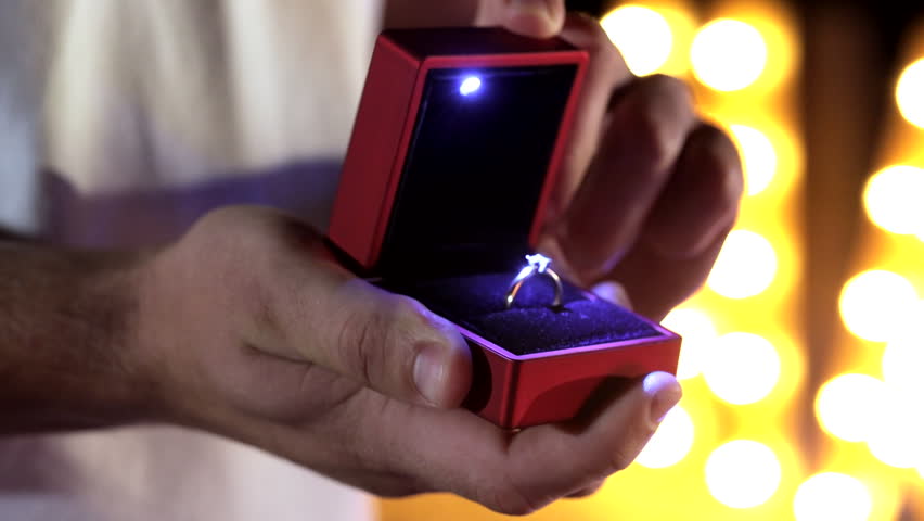 Close-up of a young man opening a red ring box with a diamond engagement ring, beautifully illuminated, set against blurred yellow lights in the evening