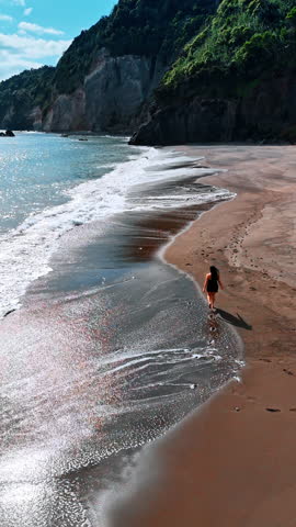 Long-haired brunette woman runs by the beautiful sandy beach at the North Atlantic Ocean shore. Rocks overgrown with lush forests are on the coast of the Azores. Top view. Vertical video.