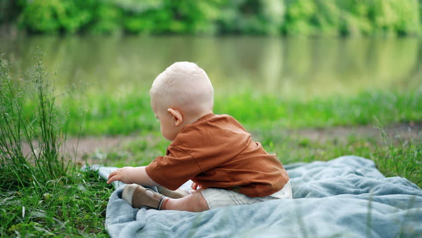 Adorable baby boy sits on the plaid in the grass. Toddler kid touches the grass with interest. Blurred backdrop.
