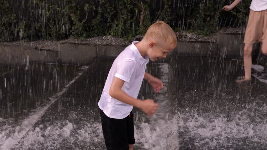 Happy children play in the fountain with a waterfall in the park in summer in hot weather, they splash and laugh. Teenagers in wet clothes are having fun in the fountain.