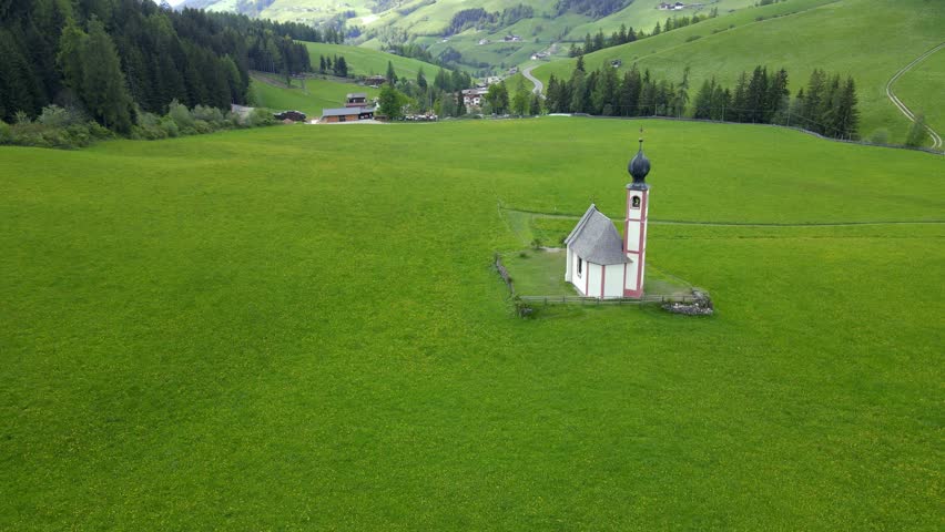 A cinematic drone glides above the Dolomites in Italy, revealing the iconic Church of St. Magdalena nestled in a green alpine valley, with dramatic rocky peaks rising majestically behind the village.