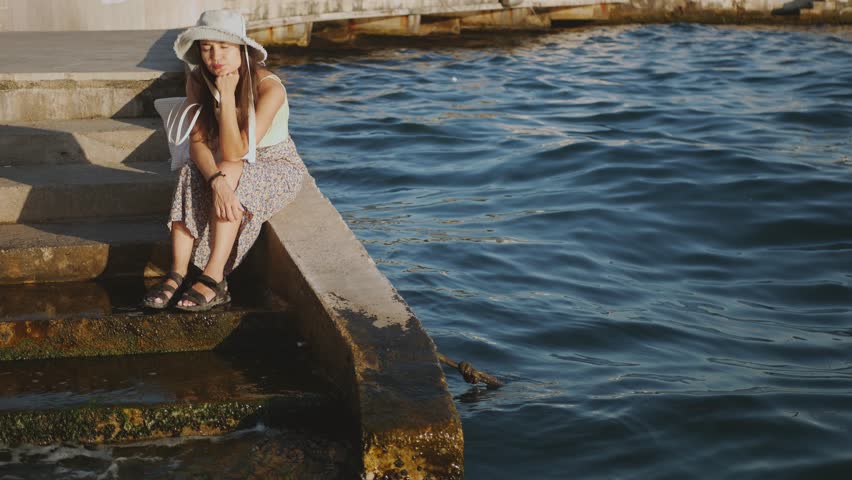 A woman in a yellow dress and white hat sits on a ledge by the water. .