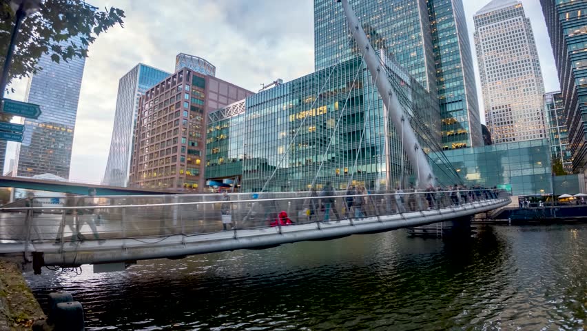 Urban concept with motion time lapse view of commuters rushing over a footbridge in front of office skyscrapers during evening, Canary Wharf, London, England