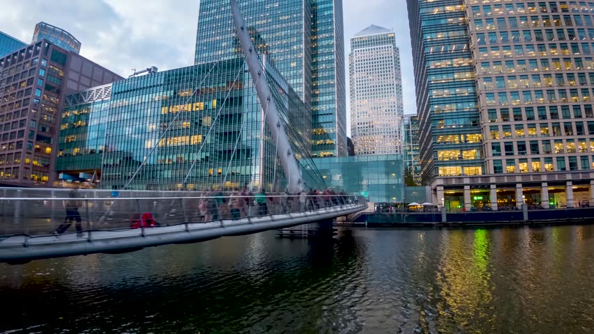 Urban concept with motion time lapse view of commuters rushing over a footbridge in front of office skyscrapers during evening, Canary Wharf, London, England