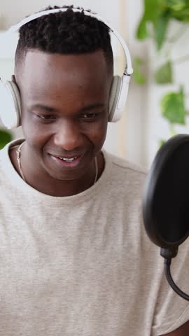 Vertical HD video of happy young black man making audio podcast from home. Content creator recording radio show using professional microphone at small broadcast studio