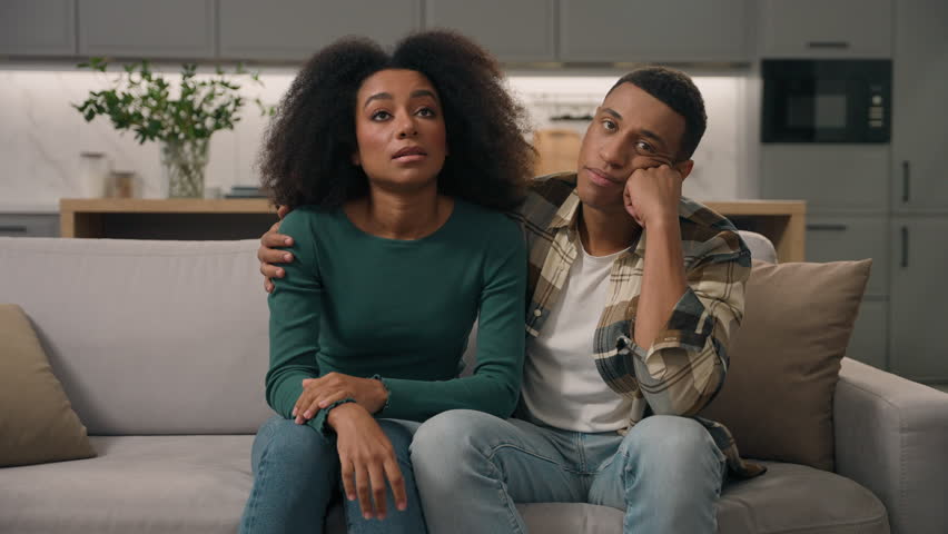 Annoyed African American ethnic exhausted young couple disturbed parents sitting sofa thinking feeling tired about noisy little boy hyperactive child misbehaving kid son playing run inside living room