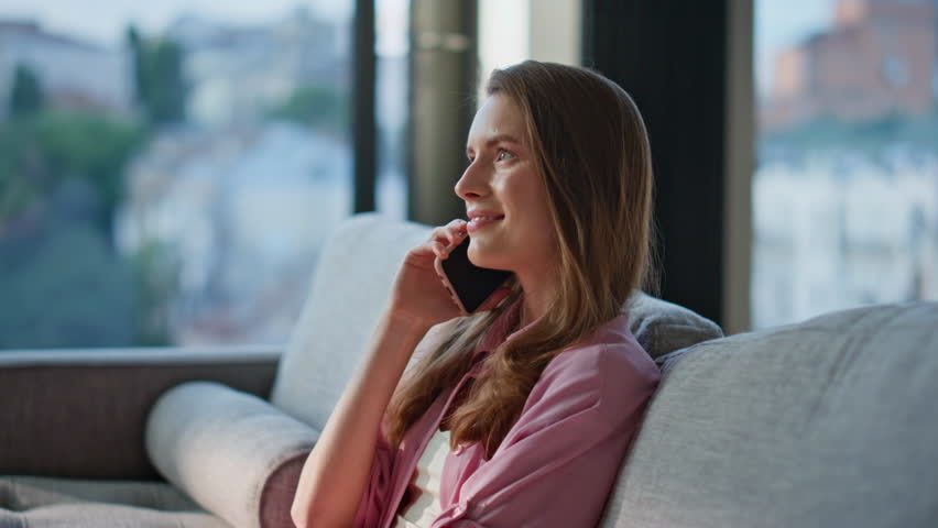Domestic girl talking cellphone sitting on apartment sofa closeup. Happy woman enjoying morning phone conversation feeling relaxed and comfortable at home couch. Cheerful lady calling in living room.
