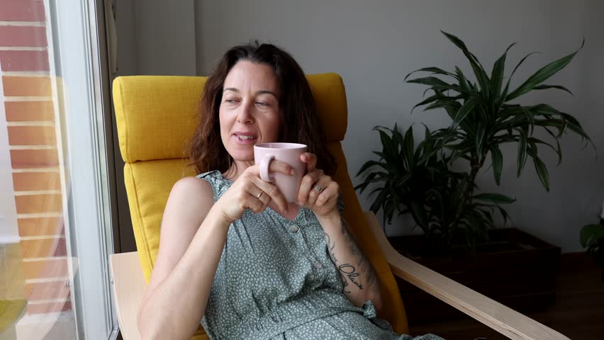 Woman is taking a moment for herself, comfortably seated in a chair by the window, holding a teacup and closing her eyes