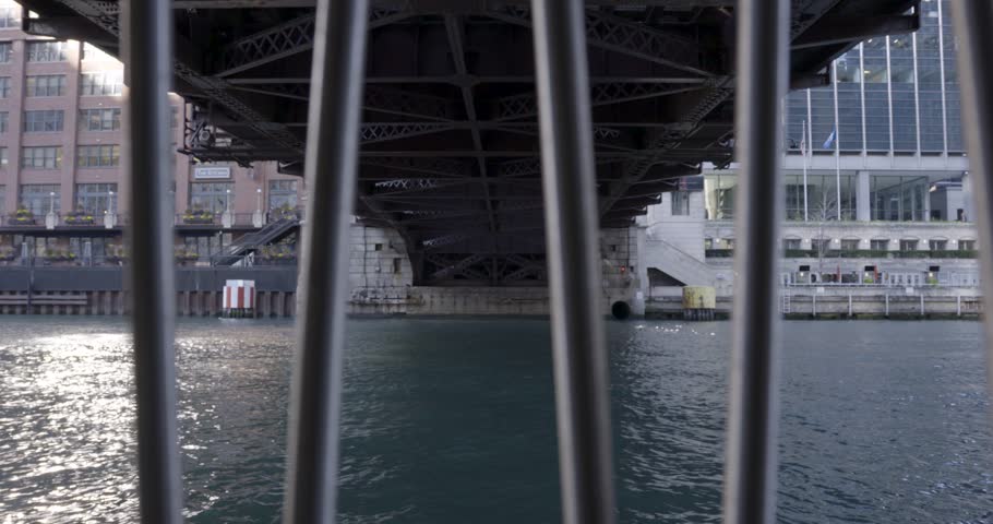 View from behind metal bars under a Chicago bridge with water reflecting nearby buildings