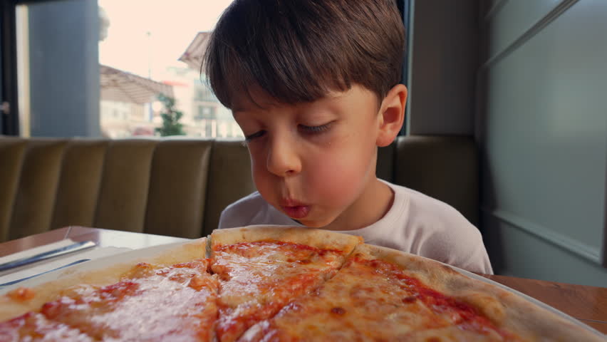 Young boy eating a large slice of pizza at a restaurant, pulling cheese with his mouth, focused on enjoying his meal in a casual and family-friendly dining setting