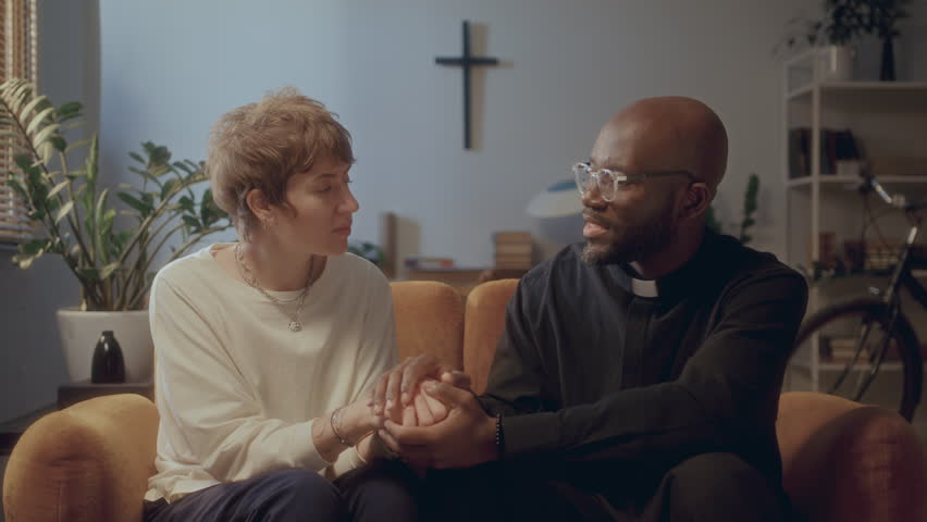Black priest in cassock and glasses holding hands of Caucasian woman and talking to her as she listening attentively to his advice on sofa at home