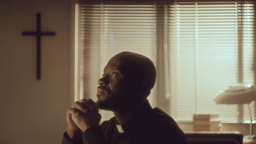African American priest in cassock looking up and saying prayer with hands clasped in warmly-lit living room with cross hanging on the wall behind