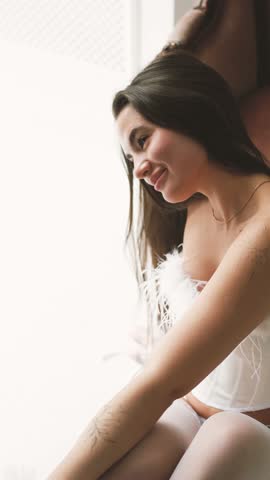 Portrait of young beautiful model posing in white lace lingerie, corset and stockings near a large window. Calm cute brunette with long hair in fashionable lingerie.