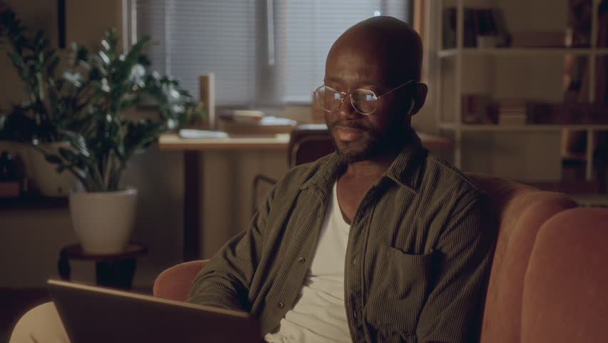 African American man wearing glasses and wireless earbuds waving and talking via video call on laptop as sitting on couch in cozy living room in the evening at home