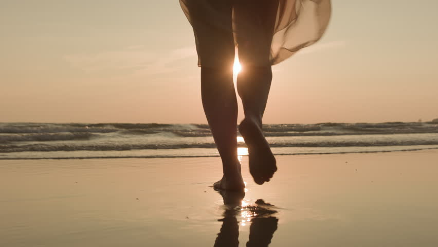 Woman feet walking barefoot in pareo towards sun on sandy beach of ocean or sea at sunset. Tourism and travel concept, still life.
