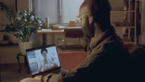 Black man wearing glasses and wireless earphones having video call on his laptop with wife or girlfriend while sitting on sofa in cozy, softly-lit room with plants and desk in the background - Powered by Shutterstock - Get 15% off with code: PIKWIZARD15
