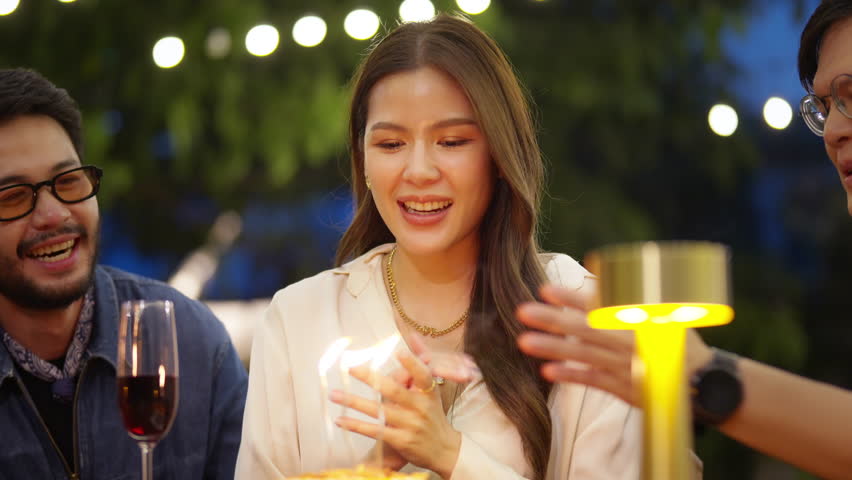 4K Group of Happy Asian millennial people friends enjoy and fun celebration birthday party at dinner party the garden at restaurant. Beautiful woman excited with birthday cake and blowing candle.