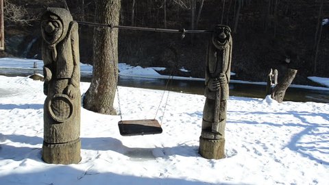 carved wooden rural craft design empty swing moving on snow covered river bank and water flow in winter.