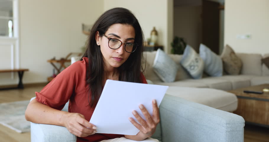 Young woman wear glasses reading document with concerned expression on her face, gesturing feels confusion or frustration, check monthly expenses, utility bills, notice from bank, bad news, troubles