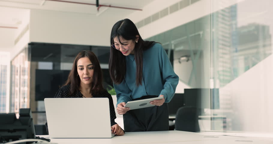 Two women colleagues collaborating on joint project in modern coworking office, look at laptop screen, engaged in discussion, reviewing documents, working on online presentation, take part in teamwork