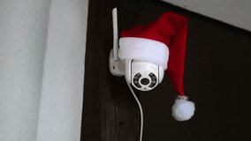 White CCTV camera in red Santa Claus hat rotates and records moving objects. Outdoor video surveillance. Security system. Web cam. Web street control. Distance watch. Smart tech. Footage. Copy space. - Powered by Shutterstock - Get 15% off with code: PIKWIZARD15