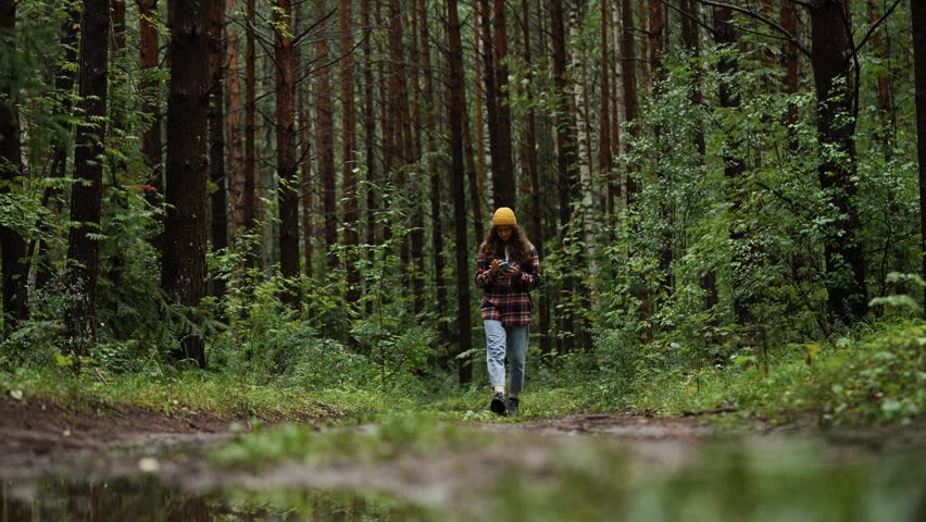 Young woman walking along a forest path uses her smartphone. Dressed in casual clothing and a beanie, she enjoys the beauty of nature while staying connected