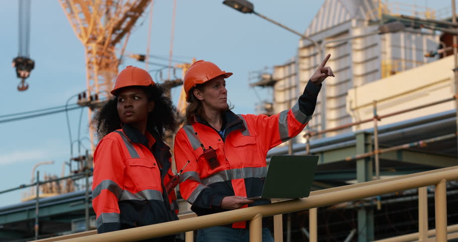 A diverse females Construction workers wearing safety helmets at an active building site with industrial equipment and structures in the background