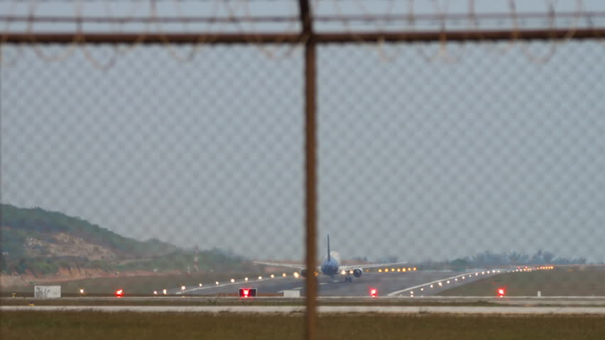 Airliner taking off and gaining altitude, rear view, long shot. Retracting, closing the landing gear. Footage of a plane behind an airport fence. Runway glowing with landing lights