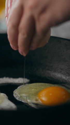 A hand cracks a fresh egg into a heated skillet, allowing the yolk to fall onto the sizzling surface, set in a cozy kitchen with natural light filling the space