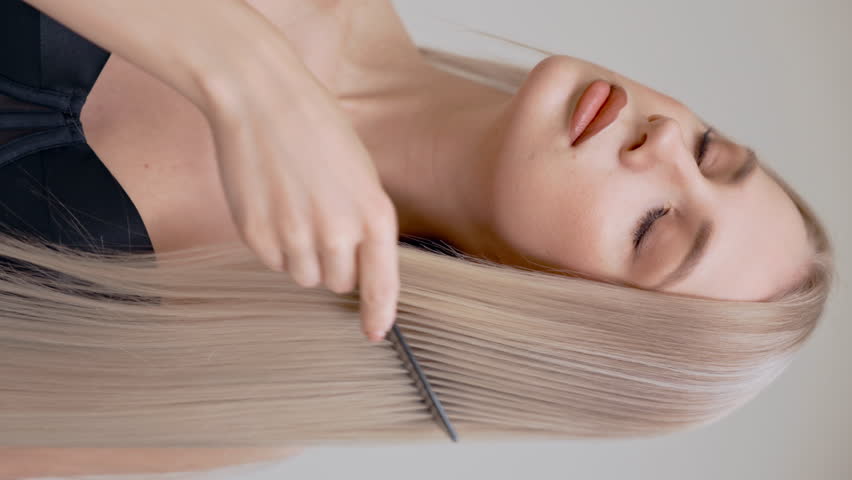 A blonde girl combs her chic long straight hair in the studio on a light background.