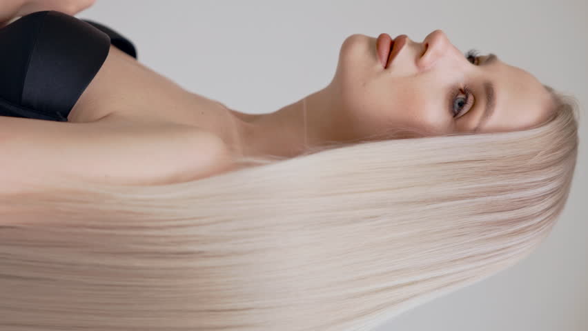 A blonde girl with gorgeous long straight hair poses in the studio on a light background. Vertical video.