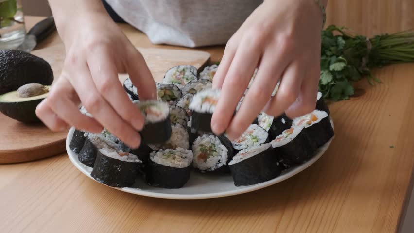 Women make sushi rolls at home from fresh ingredients like avocados, nori, rice, and vegetables on a wooden table