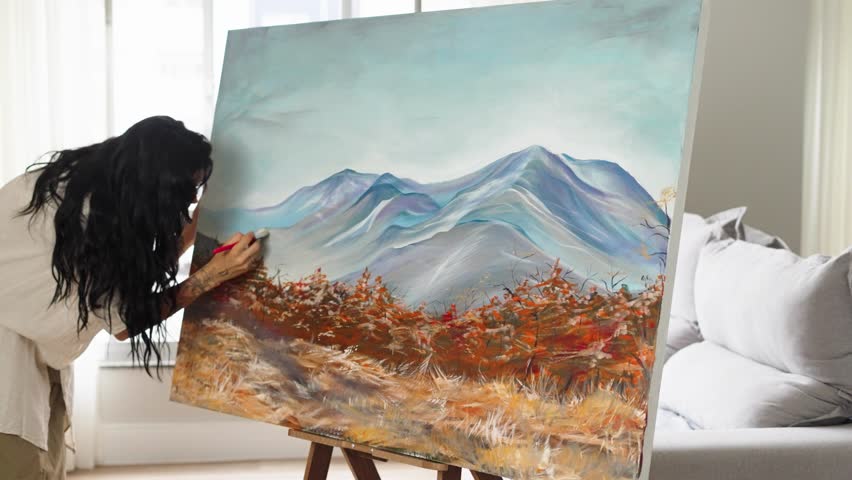 An artist applies colorful acrylic paint to a canvas with a brush. A young woman artist works in her workshop. Nature painting, contemporary art.
