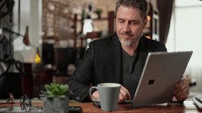 Older man sitting at desk in dark cosy room working on tablet computer in home office. Mature age, middle age, mid adult casual man in 50s, confident happy smiling. - Powered by Shutterstock - Get 15% off with code: PIKWIZARD15