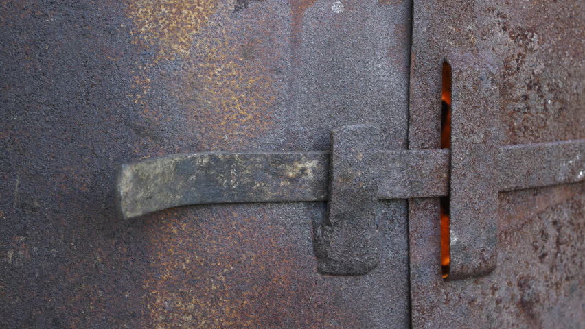 An old, rusty furnace made from a repurposed metal barrel, weathered by time and exposure, emits a warm, glowing fire from within. The flames flicker through small openings, casting an orange light.