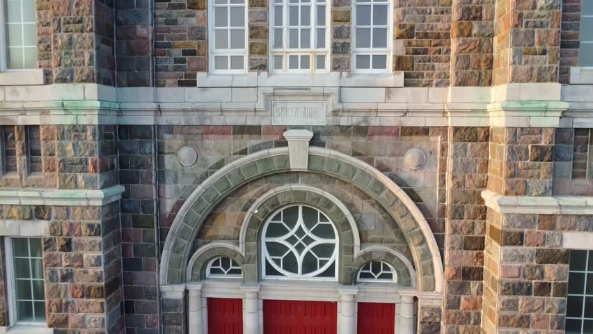 Drone video tracking up the brick and window facade of a church to a golden statue of St. Anne and her daughter Mary placed on a red roof in the center of a double bell tower. Sainte-Anne-des-Monts, Quebec, Canada.