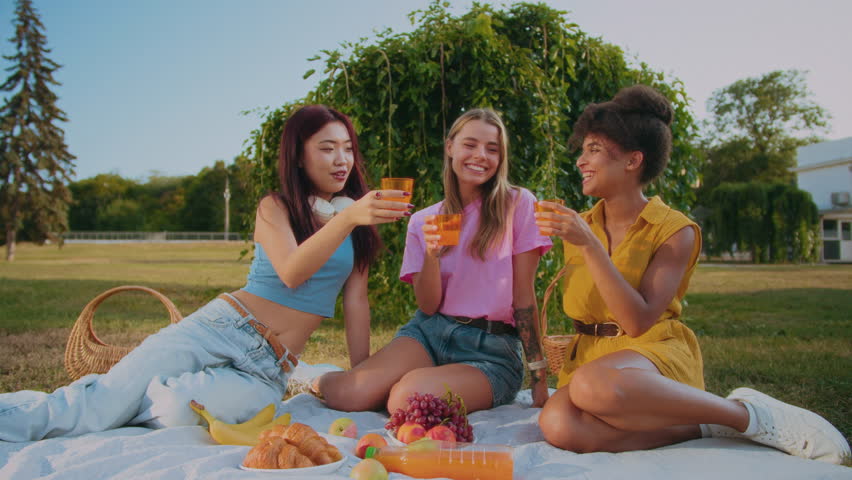 Asian, caucasian and african american women cheering with glasses of orange juice while having weekend brunch. Thirsty ladies enjoying refreshing liquid for resting from shining sun and laughing.