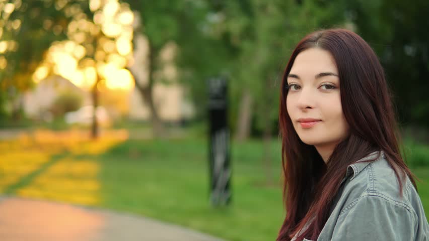 Portrait of attractive young adult Caucasian lady with long dark hair sitting in the park and looking at camera. Pretty woman is resting outdoors and smiling. Walking in town in warm weather theme