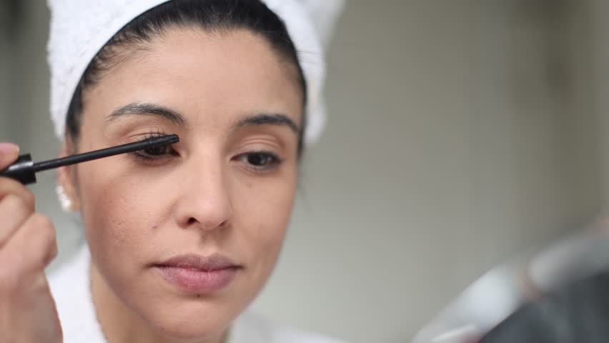 Applying mascara to eyelashes, a woman in a bathrobe and towel enhances her look while gazing into the bathroom mirror