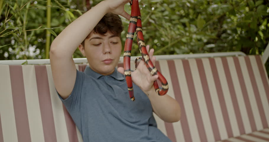 A teenage girl sits on a bench outdoors, smiling as she plays with her pet snake.