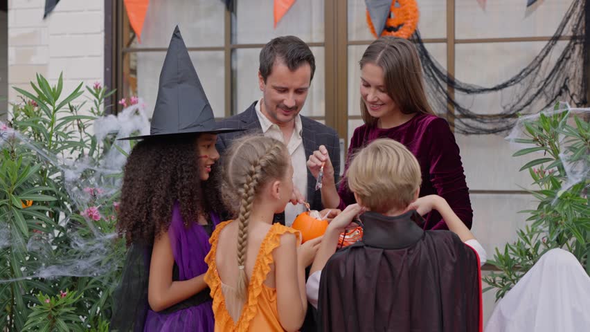 Multiracial family celebrating Halloween party together in house. Group of children dressed in Halloween costumes holding trick-or-treat pumpkin baskets while receiving candies during festival at home