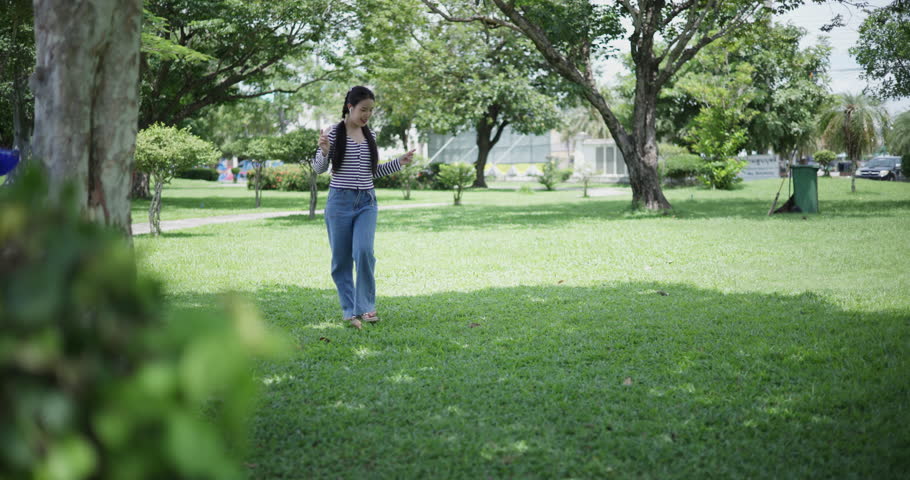 Handheld shot, Happy young woman enjoys a leisurely stroll among lush green trees in a park, wearing wireless headphones and listening to music. Weekend relaxation time