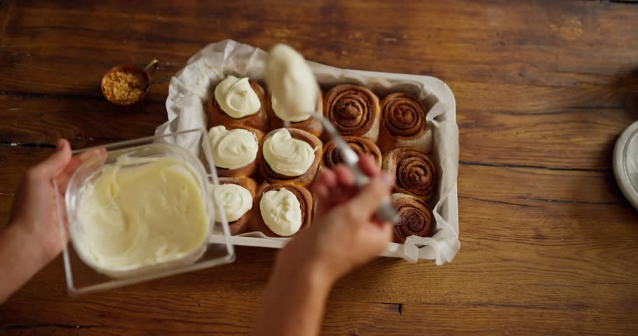 Top view of a girl baker putting white sweet cream on a prepared cinnabon cake while working in the bakery