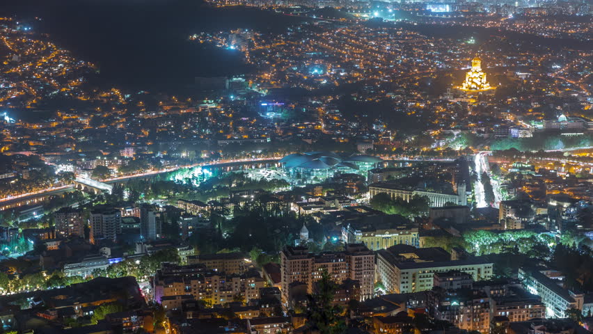 Illuminated Tsminda Sameba Cathedral or Holy Trinity Cathedral of Tbilisi and Public Service Hall aerial night timelapse. The main cathedral in Tbilisi, Georgia. Houses on background