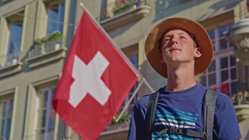 Young guy student tourist with backpack and hat in the center of the old city of Bern watching architecture, buildings around on the background of the Swiss flag. Switzerland