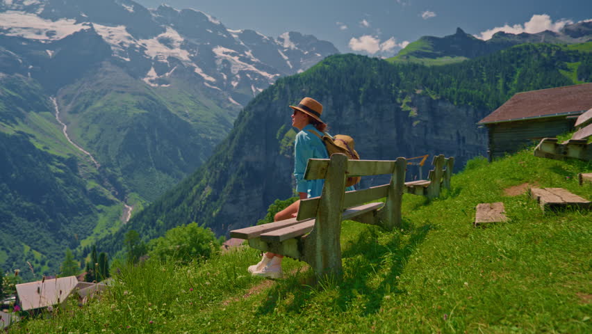 A woman tourist with a backpack sits on a bench in a very picturesque village in the Swiss Alps in the summer, looks around enjoying the beauty against the backdrop of beautiful mountains. Switzerland