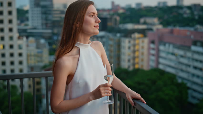 Elegant dressed woman standing on balcony enjoying peaceful evening with champagne closeup. Chic lady leaning on terrace railings holding wine glass. Gorgeous brunette looking camera at city dusk.