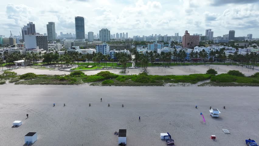 South Beach shoreline with beachgoers and umbrellas. Miami skyline in the distance with palm trees lining the coast. Aerial view of the bustling beachfront and nearby cityscape.