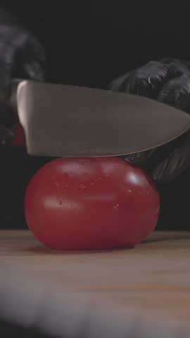 A vertical slow-motion footage of hands in gloves cutting tomato with a knife on cutting board
