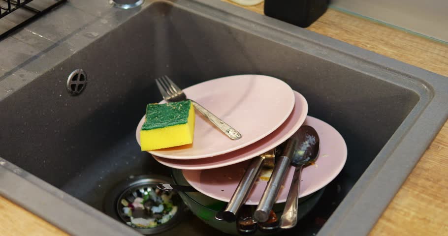 Sink filled with stack of dirty dishes with sponge on top creates smell. Food leftovers on utensils and in waste strainer harbor bacteria in kitchen